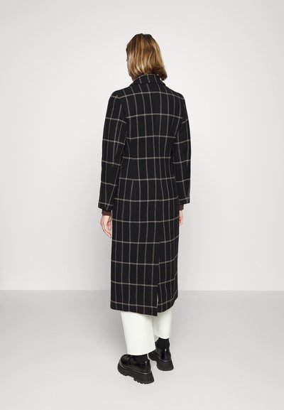 Lungo cappotto nero con un motivo a griglia beige, dotato di collo a revers e maniche lunghe. Indossato sopra pantaloni bianchi e scarpe nere spesse.