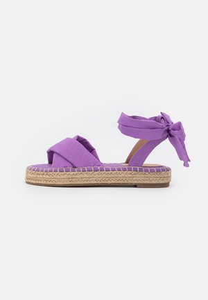 Sandale plate avec une tige en tissu violet croisée et un lacet à la cheville, dotée d'une semelle espadrille en jute tissée.