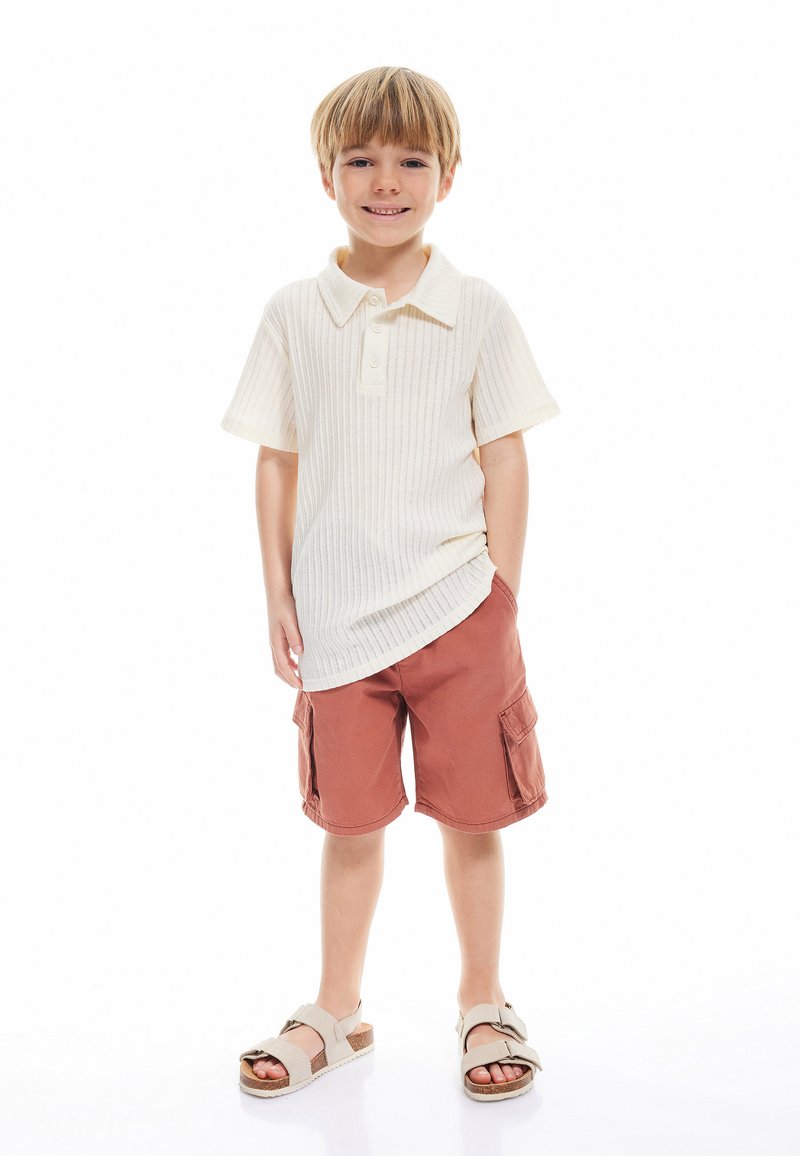Camisa polo en un color crema claro con textura de canalé, combinada con pantalones cortos cargo en color óxido y sandalias beige con un diseño de tiras.
