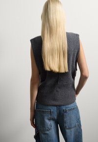 Persona con capelli biondi di spalle, che indossa una maglietta senza maniche grigio scuro e jeans di denim blu con grandi tasche posteriori.