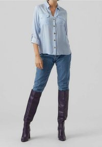 Femme portant une chemise à rayures bleues et blanches, un jean bleu et des bottes en cuir violet foncé montantes, se tenant devant un fond clair.