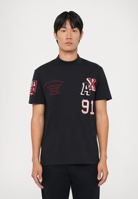T-shirt nera a maniche corte con accenti rossi ricamati, presenta i design "AX" e "91" sul davanti e un colletto alto.