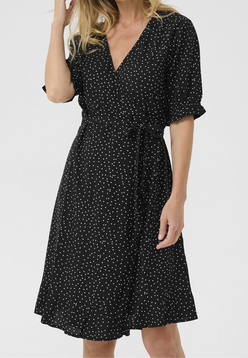 Femme portant une robe portefeuille noire à pois blancs, manches courtes bouffantes et ceinture à la taille, debout devant un fond uni.