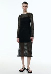 Schwarzes Netzkleid mit langen Ärmeln, das ein geometrisches Muster aufweist. Das Design ist semi-transparent, reicht bis zur Mitte der Wade und hat ein eng anliegendes Oberteil.