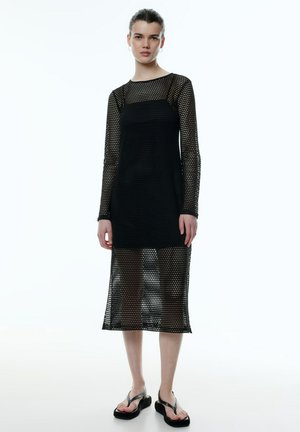 Schwarzes Netzkleid mit langen Ärmeln, das ein geometrisches Muster aufweist. Das Design ist semi-transparent, reicht bis zur Mitte der Wade und hat ein eng anliegendes Oberteil.