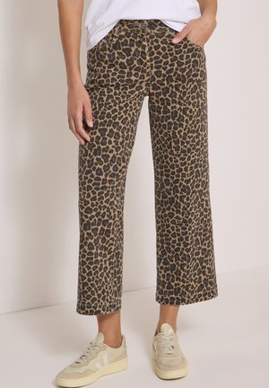 Personne portant un pantalon large imprimé léopard marron et noir et des baskets écrues, debout devant un fond clair uni.