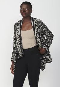 Schwarz-weißer gemusterter Blazer mit lockerer Passform. Aus gestricktem Material gefertigt, mit detaillierten geometrischen Designs und offenem Vorderteil. Kombiniert mit schwarzen, strukturierten Hosen.