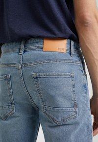 Světle modré džíny z denimu s zadními kapsami a hnědou koženou visačkou Hugo Boss, nošené s tmavě modrou košilí.