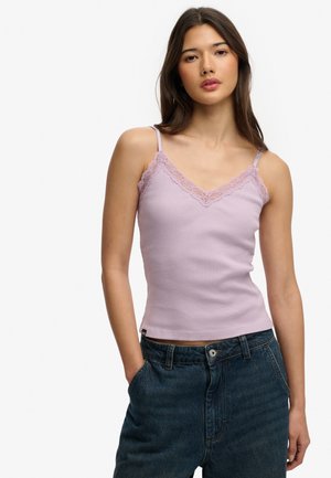 Ung kvinde iført en lys lilla top med blonder og højttaljede mørkeblå jeans, stående med hænderne i lommerne på hvid baggrund.