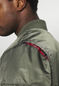 Gros plan d'une personne portant une veste en satin verte avec un détail visible en tissu à carreaux rouges et noirs à la couture de l'épaule.