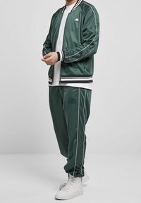 Grüner Satin-Trainingsanzug mit einer Bomberjacke, die schwarze und weiße Streifenakzente aufweist, kombiniert mit passenden Jogginghosen und weißen High-Top-Sneakers.