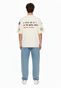 Overdimensioneret creme t-shirt med grafiske tryk på ærmerne og ryggen, der indeholder farverige tekster og kranietegninger. Kombineret med lyseblå løse jeans.