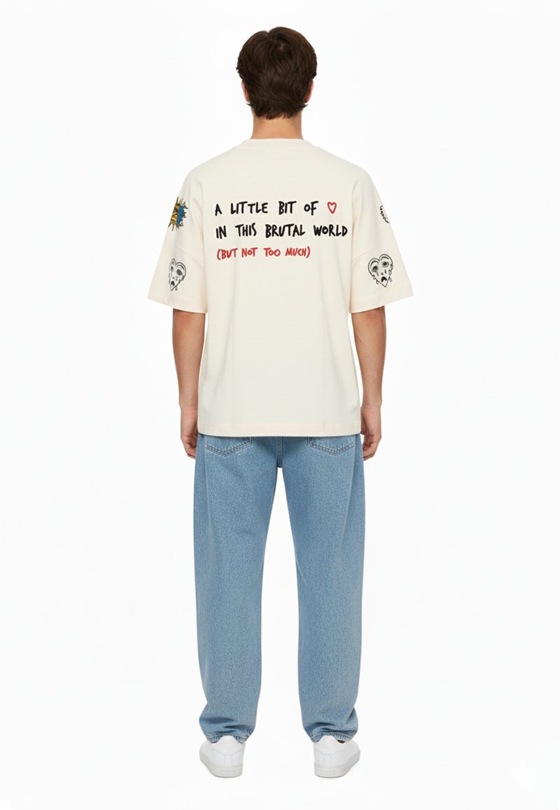 Overdimensioneret creme t-shirt med grafiske tryk på ærmerne og ryggen, der indeholder farverige tekster og kranietegninger. Kombineret med lyseblå løse jeans.