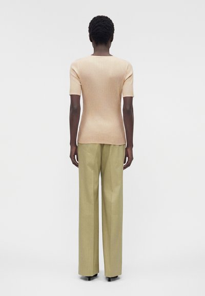 Άτομο που στέκεται με την πλάτη στραμμένη, φοράει ένα μπεζ ριπ κοντομάνικο τοπ και άνετο ανοιχτοπράσινο παντελόνι σε απλό φόντο.