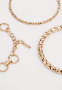 Drei goldfarbene Halsketten: eine gedrehte Seil-Halskette, eine glatte zylindrische Halskette und eine strukturierte Gliederkette mit einem kleinen Anhänger.