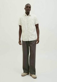 Camicia bianca a maniche corte con un motivo floreale, abbinata a pantaloni a gamba larga grigio scuro. Il modello è in piedi su uno sfondo neutro.