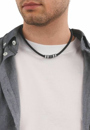 Primo piano di un uomo che indossa una collana di pelle intrecciata nera con perline cilindriche argentate, una maglietta bianca e una camicia grigia sbottonata.