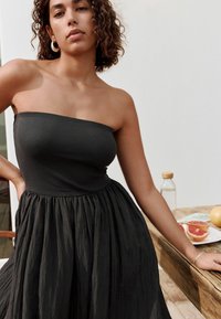 Robe noire sans manches avec un corsage ajusté à côtes et une jupe fluide et texturée. Cheveux bouclés, boucles d'oreilles créoles et table en bois avec de la nourriture en arrière-plan.