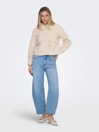 Kvinde iført en beige cropped jakke, lyseblå vidde-ben jeans og beige sko med lav hæl, stående med den ene hånd på hoften.