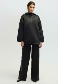 Pullover nero e oversized con cappuccio, fronte mezza zip e maniche lunghe, abbinato a pantaloni neri a gamba larga con righe bianche laterali.