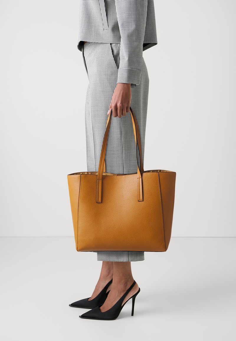 Personne portant un costume à carreaux noir et blanc, tenant un grand sac cabas en cuir marron clair, portant des escarpins noirs pointus.