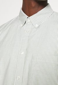 Gros plan d'une personne portant une chemise à rayures verticales vert clair et blanc, avec un col boutonné et une poche poitrine.