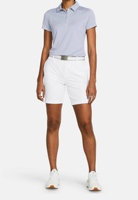 Osoba w jasnoniebieskiej koszulce polo z krótkim rękawem, białych szortach z białym paskiem i białych trampkach, stojąca na jednolitym tle.