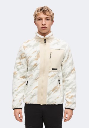 Persona joven con chaqueta de forro polar beige y blanca con estampado, con bolsillos con cremallera y pantalones negros, sobre un fondo claro y liso.