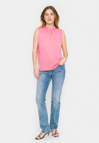 Top rosa senza maniche con scollatura plissettata, abbinato a jeans di denim blu chiaro e sandali neri con cinturini, in piedi su uno sfondo bianco.