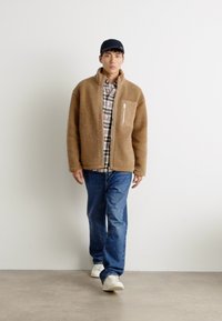 Brun fleecejakke med en sidelomme, båret over en ternet skjorte, parret med blå jeans og hvide sneakers, mod en ensfarvet baggrund.