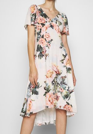 Femme portant une robe blanche à motifs floraux rose et vert, manches courtes volantées et ourlet asymétrique.