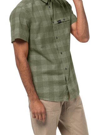 Mężczyzna w zielonej koszuli w kratę z krótkim rękawem i beżowych spodniach, z jedną ręką uniesioną przy twarzy.