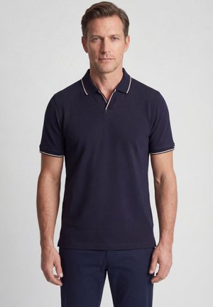 Man staat met het gezicht naar voren en draagt een marineblauw poloshirt met korte mouwen en witte biezen op de kraag en mouwen.