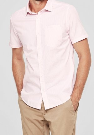 Man wearing a light pink short-sleeve button-up shirt with a chest pocket and beige pants, hands in pockets, against a plain background.