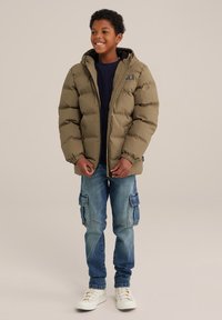 Verenpuhkuva takki oliivinvihreänä hupulla, jossa on joustavat hihansuut ja logopatch. Yhdistettynä sinisiin cargo-farkkuihin ja valkoisiin lenkkareihin.