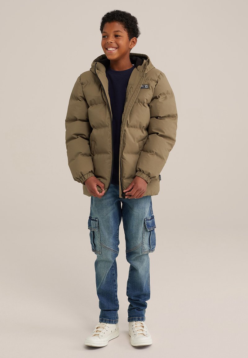 Verenpuhkuva takki oliivinvihreänä hupulla, jossa on joustavat hihansuut ja logopatch. Yhdistettynä sinisiin cargo-farkkuihin ja valkoisiin lenkkareihin.