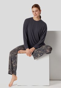 Tmavě šedá dlouhá rukávová košile s volným střihem, spárovaná s vzorovanými kalhotami s květinovým designem v béžových tónech. Model sedí na bloku.