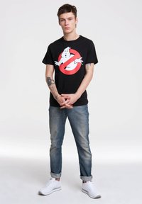 Black cotton t-shirt featuring a Ghostbusters logo in white and red. Model wears light blue denim jeans and white sneakers.