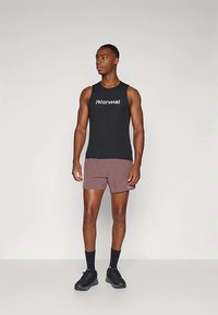 Svart ermeløs tank topp med hvit logo, kombinert med brune shorts. Modellen har på seg svarte sokker og joggesko. Enkel atletisk design.