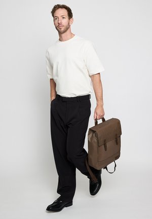 Mann trägt weißes Kurzarmhemd und schwarze Hose, hält eine braune Lederaktentasche und geht vor einem einfachen hellen Hintergrund.