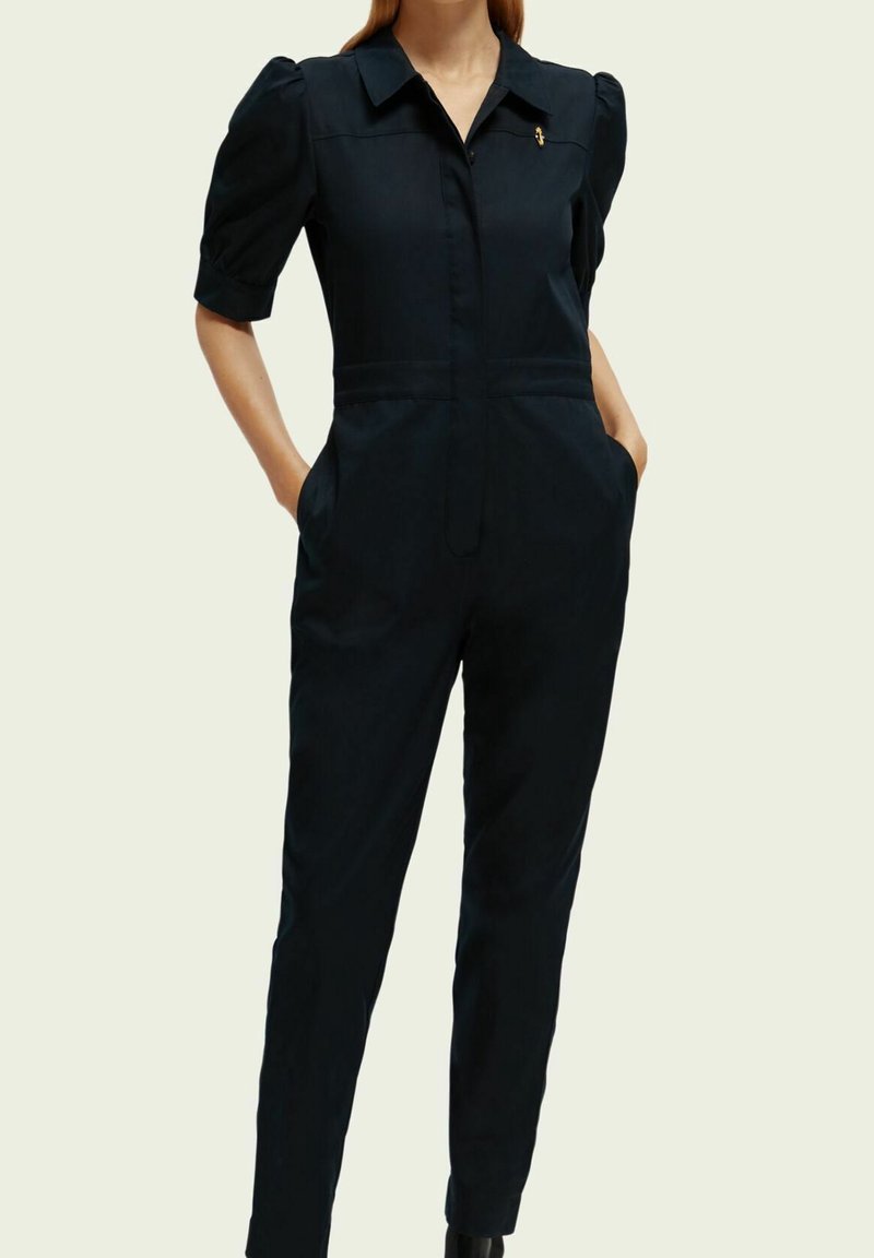 Vrouw in een zwarte jumpsuit met korte mouwen, kraag, knopen aan de voorkant, opbollende schouders, zakken en een kleine gouden spelden op de borst.