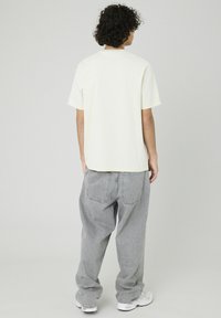 T-shirt en coton crème avec une coupe décontractée, associée à un jean en denim gris ample. La tenue présente un design minimaliste et une silhouette décontractée.