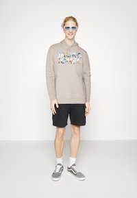 Béžová mikina s nápisom "JACK & JONES" v farebnej kvetinovej grafike, čierne cargo šortky, šedé tenisky a biele ponožky, stojí na neutrálnej pozadí.