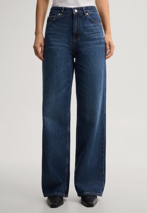 Persoon draagt donkerblauwe jeans met hoge taille en wijde pijpen en zwarte puntige schoenen, staand tegen een effen lichte achtergrond.