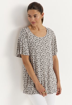 Kvinde med tilbagetrukket hår iført en hvid blomstret kortærmet top og hvide bukser, stående og kiggende let nedad.