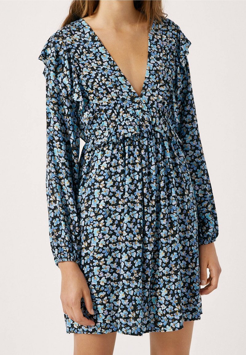 Kukkamekko pitkillä hihoilla, syvällä V-pääntiellä, mustalla pohjalla ja sinisellä kukkakuviolla. Ominaisuuksiin kuuluvat rypistyneet olkapäät ja pussitettu vyötärö rentoa istuvuutta varten.