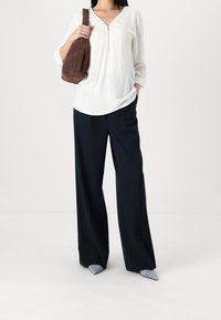 Donna con un top bianco a maniche lunghe con zip, pantaloni neri a gamba larga, scarpe grigie a punta e una borsa a tracolla marrone.