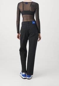 Mujer con blusa larga negra transparente, jeans negros de cintura alta con etiqueta azul de Hugo, y zapatillas blancas con detalles azules, de espaldas.