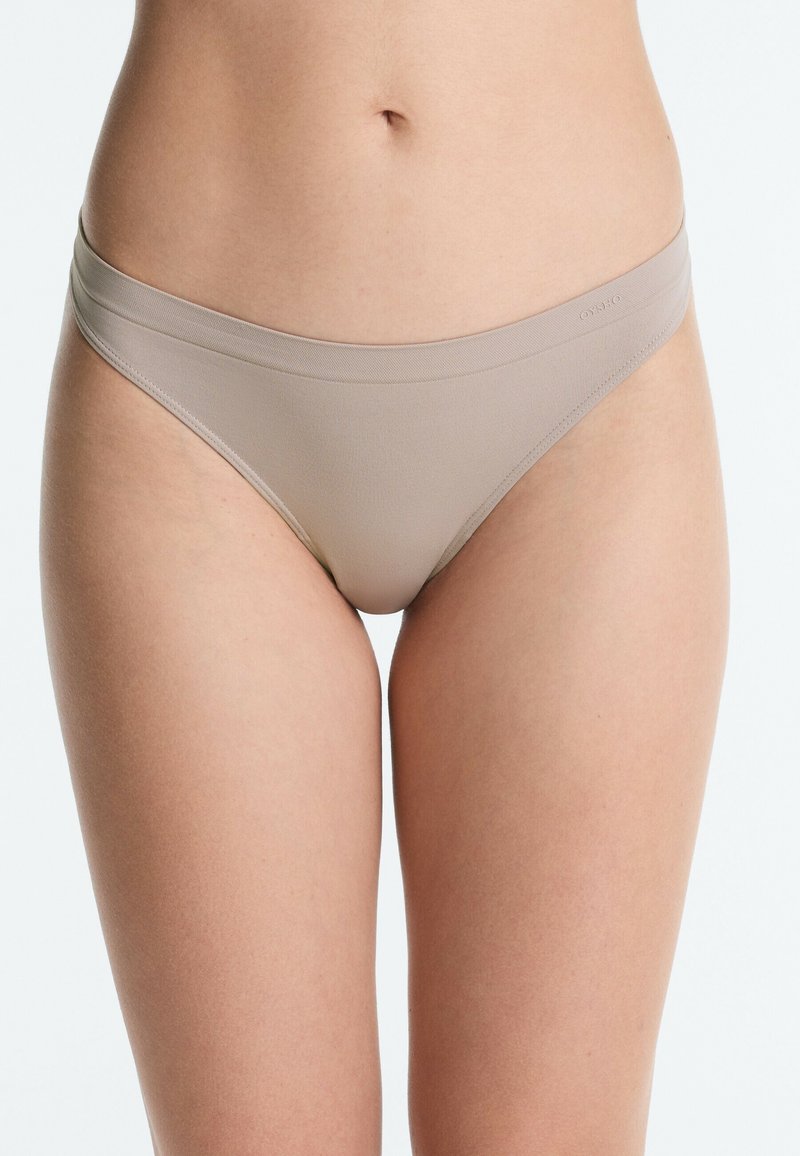 Femme portant des sous-vêtements beige sans coutures taille basse, montrant le torse du nombril aux cuisses sur un fond uni.