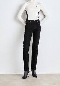 Vaqueros de denim negros combinados con una blusa de cuello alto color crema que presenta una cremallera y acentos laterales. La modelo lleva botas de tacón negro puntiagudas.